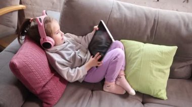 Çocuk eğlencesi ve yaşam tarzı. Küçük çocuk rahat bir şekilde kanepede uzanıyor, kulaklıklar takılı ve tablet elinde, ekrana odaklanmış. Eğlence ve evde öğrenme teknolojisi. Çocukluk kavramı