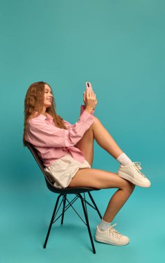 Sandalyeye yan oturmuş pembe gömlekli, beyaz etekli, deniz mavisi arkaplanlı telefon ekranına gülümseyen genç bir kadın. İnsan duyguları kavramı, yaşam tarzı, dijital iletişim, rahatlama, gençlik.