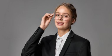 Resmi giyinmiş ve şık gözlükler takmış genç güzel bir kadının yakın plan portresi, gri stüdyo arka planına gülümseyerek poz veriyor. Mesleki yaşam tarzı, kendini ifade etme, tarz kavramı