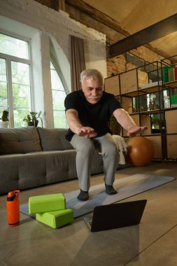 Siyah tişörtlü ve gri eşofmanlı yaşlı bir adam oturma odasında yoga minderinde çömelme egzersizi yapıyor, yerde dizüstü bilgisayara bakıyor. Yaşlılar yaşam tarzı, fitness, dijital koçluk kavramı.