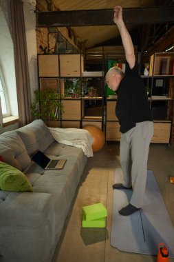 Bir kolu yukarıda yoga minderinde duran yaşlı bir adam oturma odasında egzersiz yapıyor. Laptop, yoga blokları görünür. Yaşlılar yaşam tarzı, sağlık, hareketlilik, fitness, eğitim kavramı.