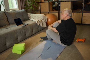 Evde sanal egzersiz yaptıktan sonra yoga minderinde oturup dizüstü bilgisayara bakan yaşlı bir adam. Yaşlılar yaşam tarzı, eğitim, bilgi, yansıma, sağlık, sağlık, sağlık.