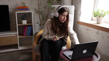 Öğretmenle çevrimiçi ders. Kulaklıkla koltukta oturan genç bir kız. İnternetteki dersi sırasında dizüstü bilgisayarla etkileşime geçip notlar alıyor. Oturma odasının içi rahat. Eğitim kavramı, e-öğrenim