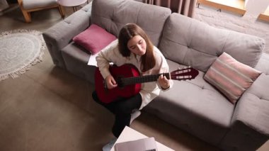 Oturma odasındaki kanepede oturan genç kızın en iyi görüntüsü, yakınlardaki masada dizüstü bilgisayarla gitar çalmak, online müzik dersleri. Öğretmenle müzik öğreniyorum. Hobi kavramı, eğitim