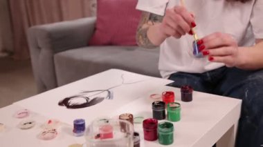 Kızıl saçlı ve dövmeli gülümseyen genç bir kadın beyaz masadaki tuvale resim çiziyor, modern oturma odasındaki yaratıcı hobisine odaklanıyor. Yaşam tarzı kavramı, sanatsal ifade, yaratıcılık, hobi.