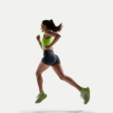 Atletik genç bir kadın beyaz arka planda öne doğru koşuyor, gri şort ve yeşil bluz giyiyor. Spor, rekabet, aktif yaşam tarzı, hız, kontrol, hareket, performans kavramı.