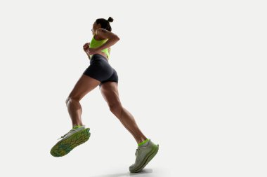 Aşağıdan yakalanan kadın sporcu, neon yeşil kıyafetiyle öne doğru koşuyor, beyaz arka planda izole edilmiş eğitim görüyor. Spor, rekabet, aktif yaşam tarzı, hareket, dayanıklılık, enerji, hız kavramı.