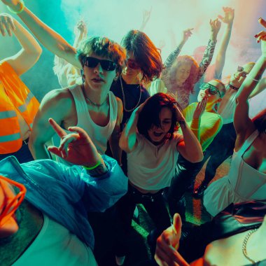 Bir grup genç bir partide ya da konserde renkli ışıklar altında enerjik bir şekilde dans ediyor. Gece hayatı kavramı, parti, gençlik, yaz, şenlik, kutlama.