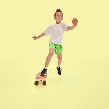Bir ayağı kaykayın üstünde atlayan komik çocuk, kolları denge için açık, sportif kıyafetler giymiş ve sarı arka planda sevinç ifadesi sergiliyor. Yaz aktiviteleri kavramı, paten eğlencesi