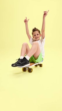 Çocuk kollarını kaldırıp kaykayın üzerinde oturuyor, geniş bir gülümsemeyle ve heyecan gösteriyor, yeşil şort ve sarı arka planda beyaz tişört giyiyor. Yaz aktiviteleri, oyun zamanı kavramı