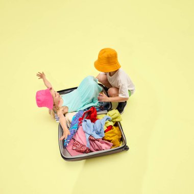 İki neşeli çocuk bavullarını topluyor, kız renkli kıyafetlerle bavulun içinde yatarken oğlan kapamasına yardım ediyor, pastel arka planda neşeli bir an. Yaz aktiviteleri, seyahat havası, dinlenme