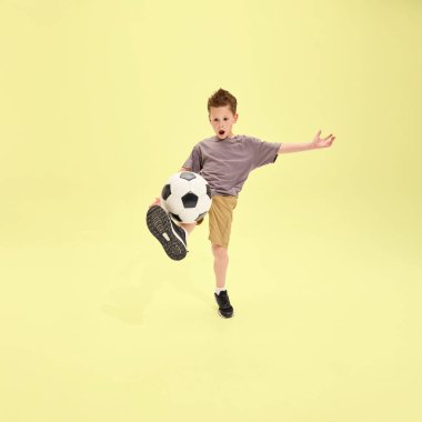 Hareketli bir çocuk futbol topunu yoğun bir şekilde tekmeliyor, gri tişört ve şort giyiyor, odaklanmış yüz, sarı arka plan. Oyun ve spor. Yaz etkinlikleri, enerji, dinlenme, tatil kavramı