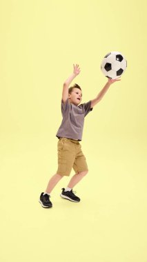 Genç bir çocuk, iki eli havada futbol topunu fırlatıyor. Stüdyo ortamında, sarı arka planda, enerjik açık hava oyunlarına karşı anlamlı bir oyuncu anı. Yaz aktiviteleri, spor, dinlenme kavramı