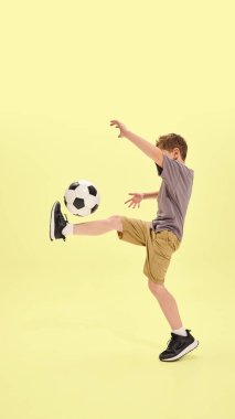 Çocuk klasik futbol hareketinde bacağını kaldırıyor, şort ve spor ayakkabı giyiyor, sarı arka plan hareket ve koordinasyona odaklanıyor. Yaz aktiviteleri, hobi, tatil kavramı