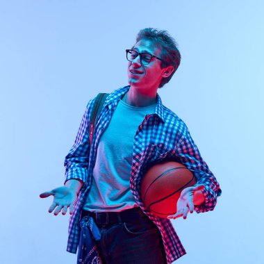 Gözlüklü genç bir çocuğun portresi, gündelik giysiler ve basketbol topu taşıyan sırt çantası, neon lambası altında eğimli arka plana karşı komik, kafası karışmış bir suratla omuz silkiyor. Spor anlayışı, iletişim