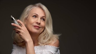 Serum şişesini tutan olgun bir kadın ve siyah arka planda neşeli bir şekilde gülümseyen parlak bir ten. Güzellik tedavisi kavramı, cilt rutini, kaldırma serumu, sağlıklı yaşlanma, estetik ameliyat