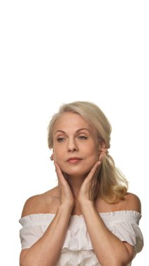 Zarif yaşlı kadın çene hattına ellerini bastırıyor, beyaz arka plana masaj yapıyor, kendinden emin yaşlanma ve günlük yüz bakımını gösteriyor. Yaşlanma karşıtı tedavi kavramı, kozmetik ürünler