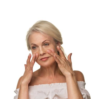 Yaşlı kadın parmak uçlarını kullanarak göz altlarındaki deriyi beyaz arka plana bastırıyor, şişkinliği azaltıyor ve cilt bakımını özgüvenle destekliyor. Yaşlanma karşıtı kavram, göz tedavisi, cilt bakımı