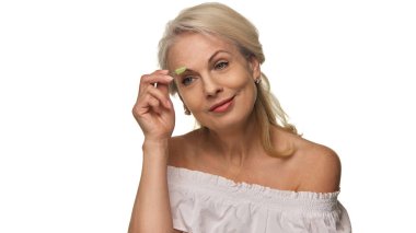 Zarif, son sınıf öğrencisi kadın kaşlarını küçük yeşil bir fırçayla şekillendirirken omuz üstü beyaz bir bluzla gülümsüyor, özgüveni ve rutini teşvik ediyor. Günlük güzellik kavramı, kozmetik