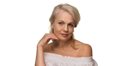 Beyaz bluzlu sarışın olgun kadın bir eliyle çenesine hafifçe dokunuyor, beyaz arka plana karşı özgüven ve cazibeyle gülümsüyor. Yaş-pozitif zarafet ve güzellik kavramı