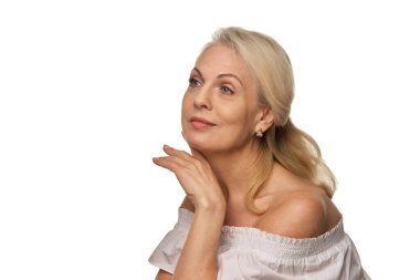 Zarif yaşlı kadın, beyaz arka plana karşı düşünceli ve ışıl ışıl bir anda nazikçe çenesinin altından elini tutan yumuşak bir gülümseme ile yukarıya bakar. Olgun güzellik anlayışı, spa tedavisi, sağlık.