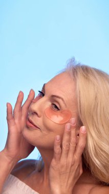 Kıdemli kadın gözlerinin altına şeftali rengi yamalar yerleştirir, gözlerini kapatır ve güzellik rutininin tadını çıkarırken hafifçe gülümser. Göz bakımı, yaşlanma karşıtı, kaldırma, kozmetik ürünleri kavramı