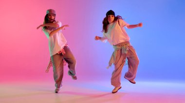 Dinamik fotoğraf. Pembe ve mavi ışıkların altında büyük beden kıyafetlerle dans eden iki kız. Gradyan stüdyo geçmişine karşı. Müzik kavramı, ritim, sokak enerjisi ve şehir atmosferi.