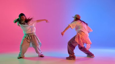 Serbest stil hip hop dansı yapan iki kız parlak pembe ve mavi ışıklar altında gradyan stüdyo arkaplanına karşı hareket ediyor. Dinamik fotoğraf. Müzik kavramı, modern hareket, moda ve sokak ritmi..