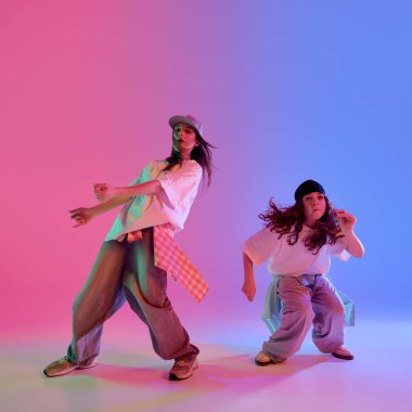 Pembe ve mavi neon ışıkları altında hiphop hareketleri yapan şık giyimli genç kızlar gradient stüdyo arka planına karşı. Müzik, ritim, tarz ve dışavurumsal hareket kavramı.