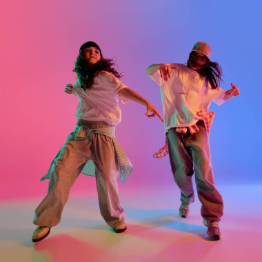 Dinamik ikili, parlak atletik genç kızlar, parlak neon ışıklarıyla dans eden sokak tarzı pembe ve mavi gradyan stüdyo arka planı. Müzik anlayışı, gençlik enerjisi ve anlamlı hareket.