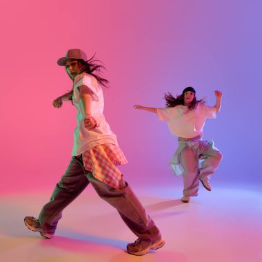 Kızlar, hareketli hip hop dansı sırasında pembe ve mavi neon ışıklarının altında gradyan stüdyo arka planına karşı hareket halindeler. Müzik, gençlik kültürü, kentsel hareket ve dinamik akış kavramı.