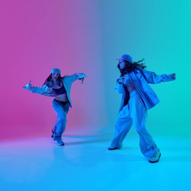 İki genç, sanatsal hip hop dansçısı, kızlar, cesur şehir moda kıyafetleri giymişler, gradyan arka planına karşı parlak bir ışık altında birbirlerine bakıyorlar. Sokak kültürü kavramı, tutum, hareket.
