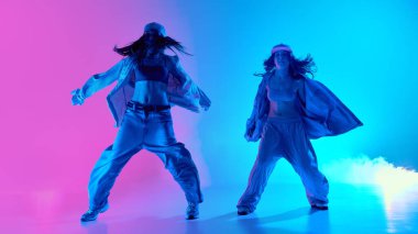 Artistik genç kızlar, serbest stil hip hop ikilisi stüdyoda turkuaz ve pembe ışıklandırma efektleri ile gradyan arka planda pratik yapıyor. Müzik kavramı, dinamik biçim, biçim ve hareket akışı.