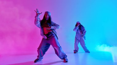 İki şehir dansçısının gradyan pembe ve cyan ışığı altında cesur hip hop hareketleri yaparken çekilmiş dinamik bir fotoğrafı. Ritim kavramı, bireysellik ve sokak modasına uygun dışavurumcu hareket.