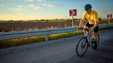 Sarı bisikletli bir adam, gün batımında kırsal yolda bisiklet sürüyor odaklanmış ifadesi ve güçlü duruşuyla. Spor, bisiklet, dayanıklılık, sağlıklı yaşam tarzı, gün batımı yolculuğu kavramı.