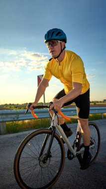 Altından ışıkta, koruyucu giysiler ve spor kıyafetlerle yokuş yukarı pedal çeviren sporcu virajlı yolda ilerliyor. Hareket bulanıklığı etkisi. Bisiklet sporu konsepti, fitness eğitimi, gün batımı, kararlılık.