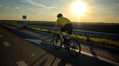Altın saat boyunca ufka doğru ilerleyen bisikletçinin arka görüntüsü, hareket bulanıklığı etkisiyle açık yol ve güneş önümüzde. Dayanıklılık kavramı, kişisel yolculuk, sağlık odağı, spor adanmışlığı.