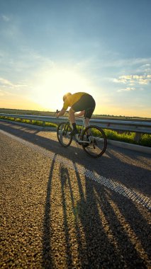 Düşük açılı çekimde bisikletçi sıcak güneşin altında uzun bir gölge yaratıyor ve hareket halindeki güçle pedal çeviriyor. Enerji, hareket, spor atmosferi, motivasyon, çaba ve gündoğumu eğitim yolculuğu kavramı.