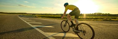Bisikletçi, arka planda parlayan güneş ve yeşil tepeler görünürken geniş asfalt şeridinin yumuşak kıvrımlarında yol alır. Pankart. Spor anlayışı, bisiklet, sağlıklı yaşam tarzı, dayanıklılık, doğa.