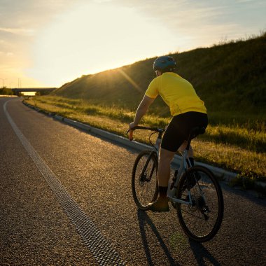 Bisikletçi, çimli yamaçlar arasındaki yolun sessiz eğrisinde uzun gölgeli, yumuşak altın ışıkla ufukta süzülür. Spor anlayışı, bisiklet, sağlıklı yaşam tarzı, dayanıklılık, doğa
