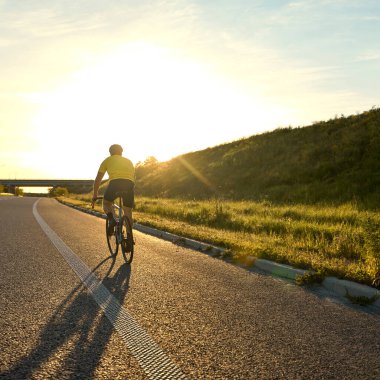 Bisikletçi parlak ufukta tek başına ilerlerken, gece ışığında çimenli yamaçların yanında, uzun ve temiz bir yolda ilerler. Spor anlayışı, bisiklet, sağlıklı yaşam tarzı, dayanıklılık, doğa.