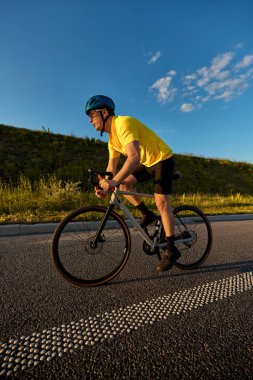 Sarı formalı odaklanmış bir adam, canlı mavi gökyüzü ve sakin yoldaki altın saat ışığı altında kendine güvenen bir duruşla yokuş yukarı gider. Spor anlayışı, bisiklet, sağlıklı yaşam tarzı, dayanıklılık, doğa.