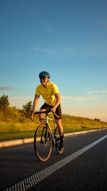 Sarı formalı bisikletçi, yeşil tepeler ve parlayan gökyüzüyle çevrili açık otoyolda yoğun günbatımı antrenmanı sırasında güven içinde ata biniyor. Spor, bisiklet, dayanıklılık, ırk kavramı,