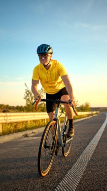 Sarı gömlek pedallı sporcu, akşam ışığı ve düz yol tarafından çerçevelenmiş solo bisiklet egzersizi sırasında odaklanma ve kararlılık ile yokuş yukarı gidiyor. Spor, bisiklet, eğitim, rekabet kavramı