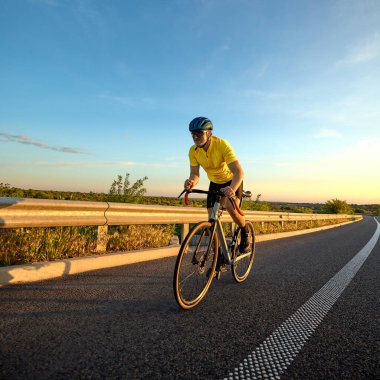 Bisikletçi, günbatımı parıltısı ve pürüzsüz kır yolu ile çevrili yoğun eğitim yolculuğunun hızlandırma safhasında öne doğru eğiliyor. Spor anlayışı, ırk hazırlığı, bisiklet, dayanıklılık, hız