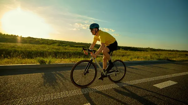Kask pedallı bir adam, gün batımında parlayan geniş yol boyunca sorunsuzca pedal çeviriyor. Dengesini koruyor ve yeşil alanların yakınında istikrarlı adımlar atıyor. Spor anlayışı, bisiklet, sağlıklı yaşam tarzı, dayanıklılık, doğa.