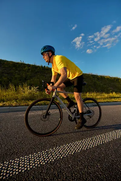 Sarı formalı odaklanmış bir adam, canlı mavi gökyüzü ve sakin yoldaki altın saat ışığı altında kendine güvenen bir duruşla yokuş yukarı gider. Spor anlayışı, bisiklet, sağlıklı yaşam tarzı, dayanıklılık, doğa.