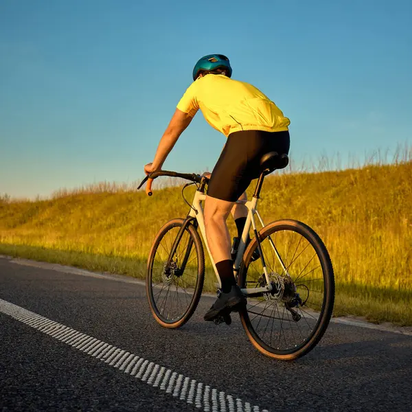Sürücünün kırsal yolda sabit bir şekilde yuvarlanışının arka görüntüsü, son kilometrelerce uzunluğundaki eğitim döngüsünü altın saatte tamamlıyor. Spor, bisiklet, rekabet, rutin, mesafe, egzersiz, motivasyon kavramı