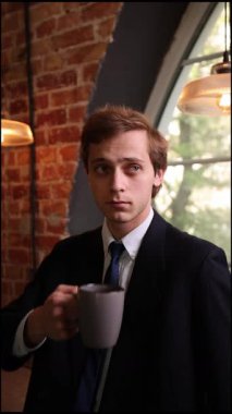 Siyah takım elbiseli şık bir adam kahve bardağını geniş bir pencereden tutuyor. Sessiz anın tadını çıkarmak için işten ara veriyor. Güneş ışığı odaklı yüzüne hafifçe parlıyor. Yaşam-iş dengesi kavramı, iş günü.