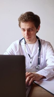 Beyaz önlüklü erkek doktor, dizüstü bilgisayarda yoğun ifadeyle oturuyor ve klinikte dijital kayıtlarla çalışıyor. Çevrimiçi danışma, tanı, iş akışı kavramı.